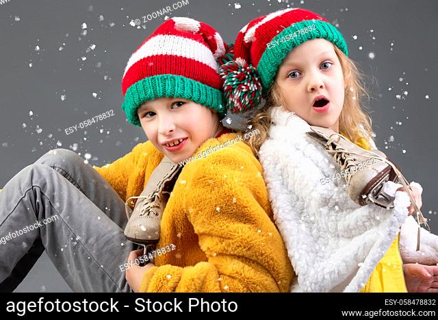 Funny little girl and boy in knitted Christmas hats and vintage ice skates sit with their backs to each other and look at the falling snow on a gray background