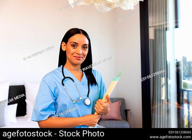 Portrait of biracial smiling female physiotherapist with report and stethoscope standing at home