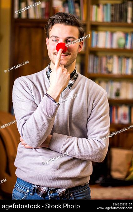 Handsome young man smiling and touching red clown nose, standing in a living room
