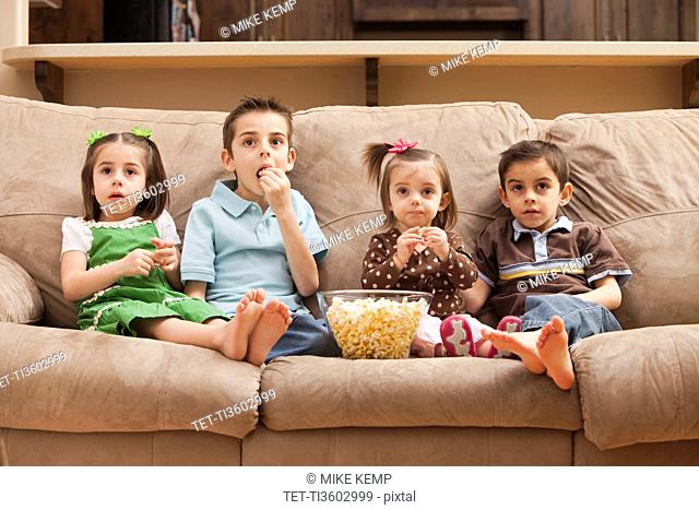 Front view of children 18-23 months, 4-5, 6-7, 8-9 sitting on couch watching tv and eating popcorn