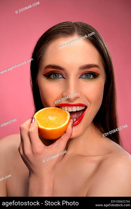 Beautiful fashion girl holding orange sliced in half in her hand biting it looking at camera. Charming joyful funny lady with red lips and long hair isolated on...
