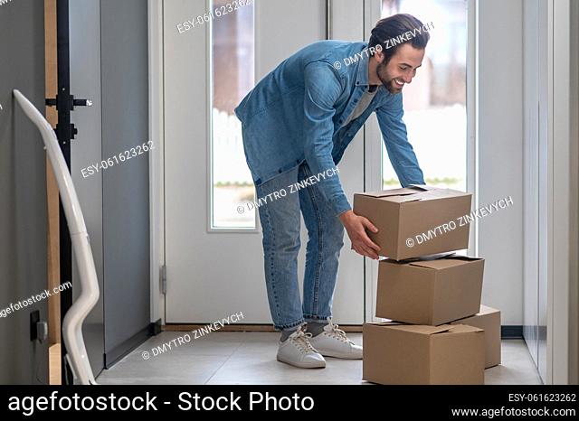 Boxes, moving. Smiling young bearded caucasian man standing bent over stacking boxes near glass door in apartment