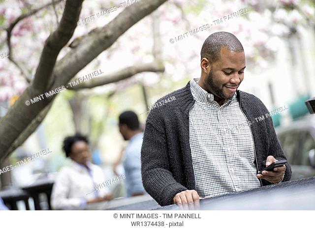 Outdoors in the city in spring. An urban lifestyle. A young man checking his phone and texting. A couple in the background