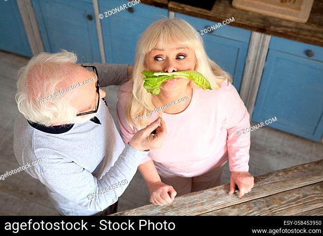 Couple of senior people having fun while making lunch. Charming old woman being silly putting lettuce on her upper lip so it looks like mustache