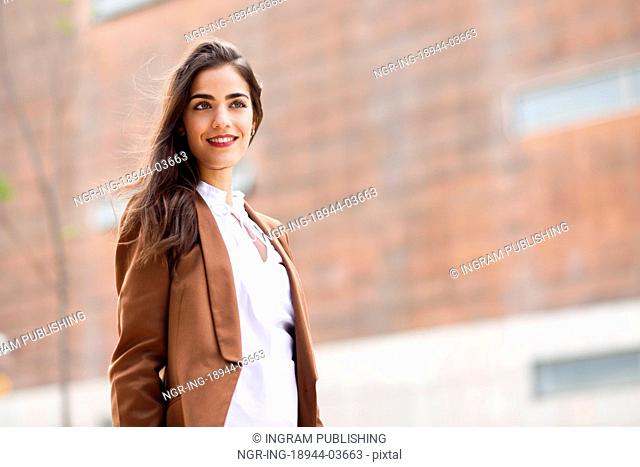 Young woman with nice hair standing outside of office building. Businesswoman wearing formal wear with wavy hairstyle. Young girl with brown jacket and trousers