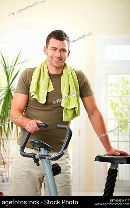 Man wearing sportswear and towel standing in living room at home with training bike, smiling