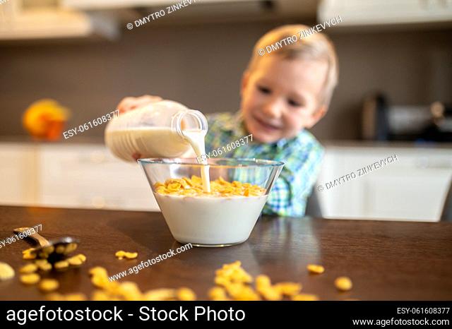 Smiling kid adding milk from a plastic bottle to dry cereals in the glass bowl