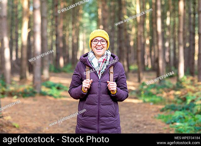 Happy woman in warm clothing holding backpack while standing in forest