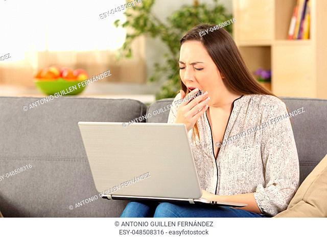 Bored woman on line yawning sitting on a sofa in the living room at home