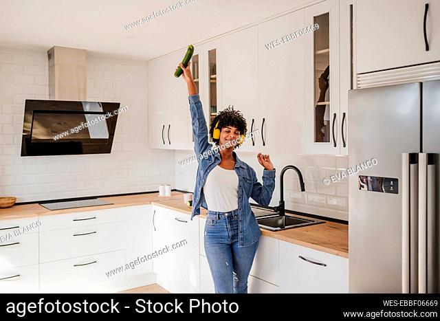 Smiling woman wearing wireless headphones dancing with zucchini in kitchen at home