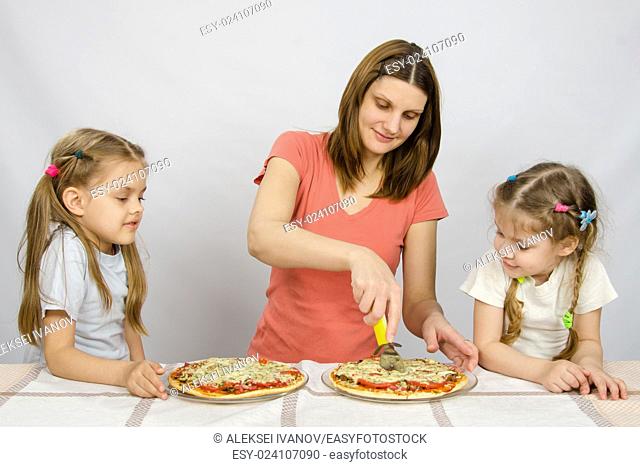 Mom cuts the pizza, and the two little girls eagerly look