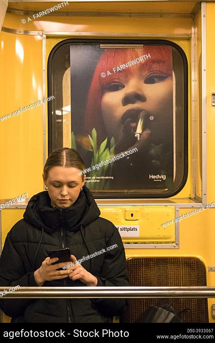 Stockholm, Sweden A young beautiful woman sits on the tunnelbana under a poster of an oriental woman applying lipstick