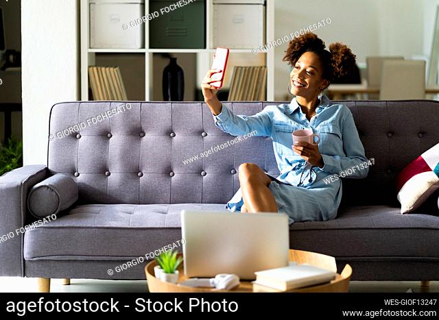 Smiling woman sitting with coffee cup while taking selfie through smart phone at home