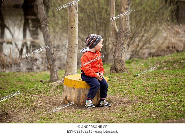 Little boy sitting on a stump in the spring park. Baby boy in orange ja?ket, outdoors