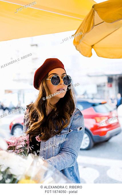 Young woman in beret and sunglasses shopping for flowers on urban sidewalk