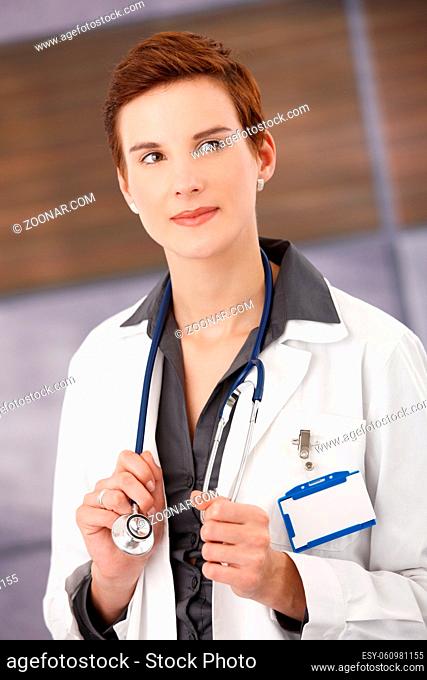 Smiling female physician standing in office wearing smock, holding stethoscope