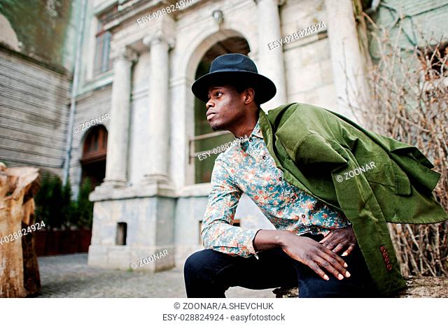 Fashion portrait of black african american man on green velvet jacket at his shoulders and black hat, sitting at felled tree background old vintage house with...