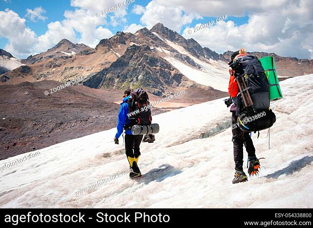Two tourists, a man and a woman with backpacks and crampons on their feet walk along the glacier against the background of the mountains of the sky and clouds