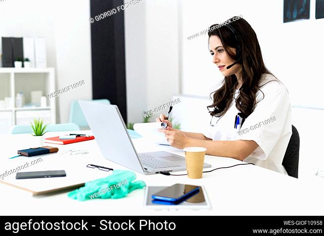 Young female doctor writing on notepad while consulting online through laptop in clinic