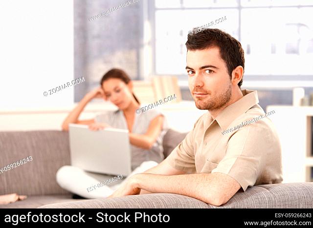 Young man sitting on sofa at home, woman using laptop in the background