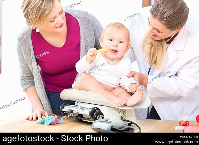Female pediatric care specialist smiling while measuring the weight of a cute and healthy baby girl held by her mother during routine medical check-up