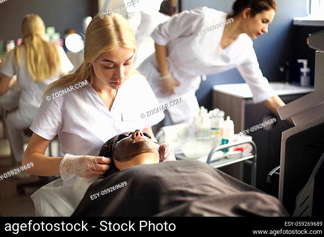 Female cosmetologist applying black mask on the face of a beautiful woman for carbon peel
