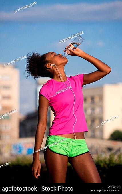 Young beautiful african american woman with headphones drinking water after jogging in the city