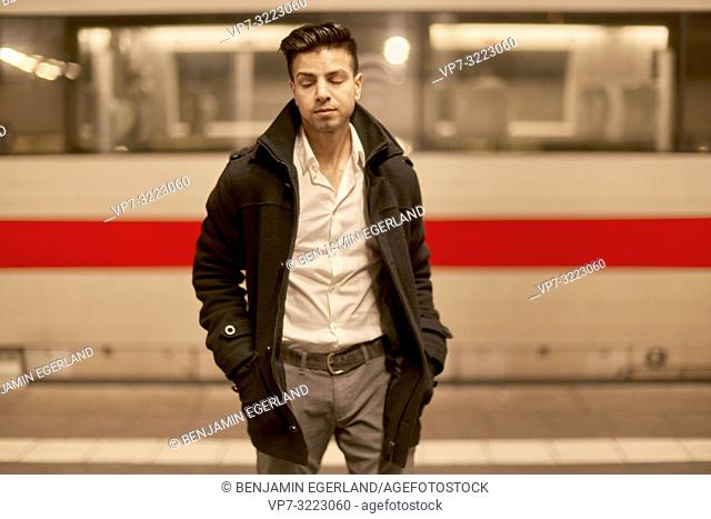 young tired man in front of train at train station with closed eyes, Afghan ethnicity, in Munich, Germany