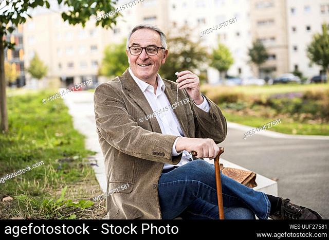 Smiling senior man holding in-ear headphones sitting with walking cane
