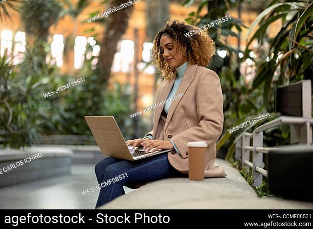 Businesswoman using laptop sitting with disposable cup on bench