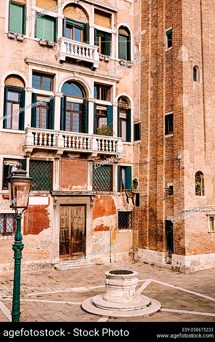 An old street well in the square in front of a brick house. There are many Venetian-style windows on the facade of the building