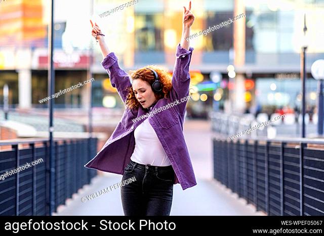 Cheerful woman with arms raised dancing while listening music through headphones on bridge