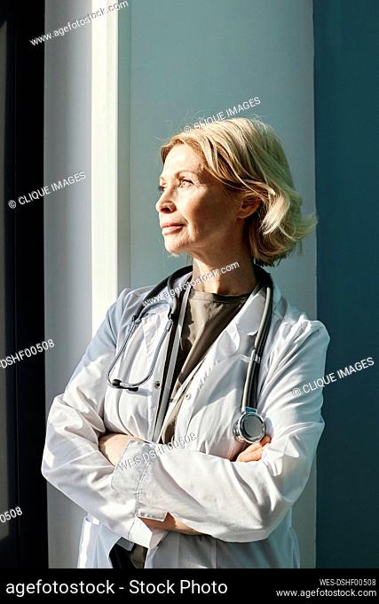 Thoughtful doctor standing with arms crossed by wall