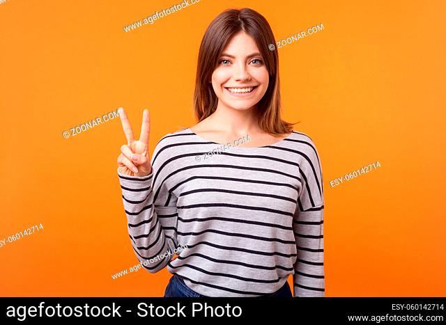 Portrait of happy charming woman with brown hair in long sleeve striped shirt smiling looking at camera and showing peace or victory sign with fingers