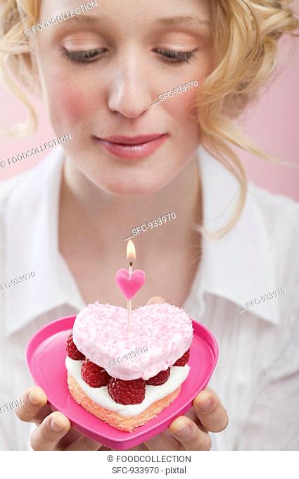 Young woman holding raspberry cake with candle in heart-shaped dish