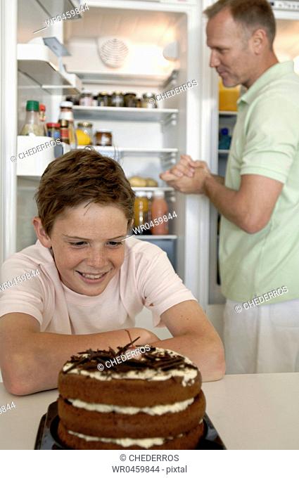 Boy looking at a cake with his father standing behind him in front of an open refrigerator