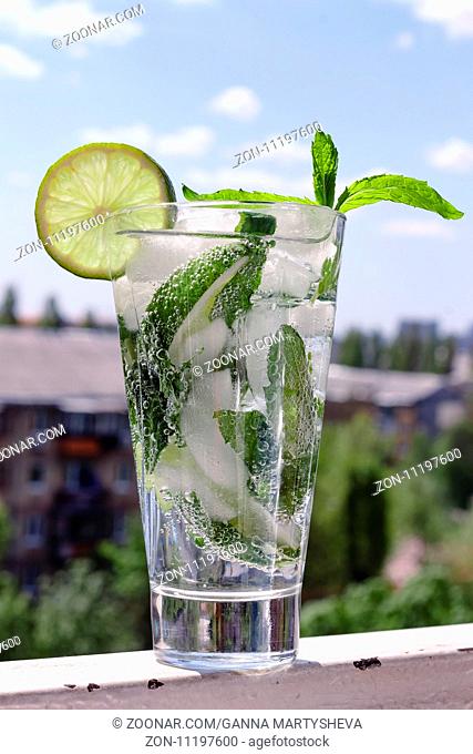 Mojito in a glass on a background of a modern city and blue sky with clouds. Closeup. Alcoholic or non-alcoholic cocktail