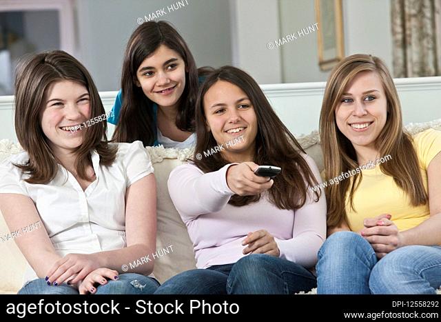 Four teenage girls sitting on a couch at home watching TV and changing the channel with the remote control