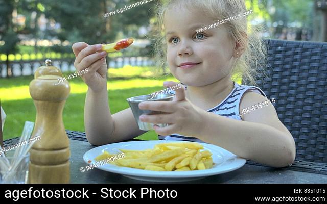 Little girl eat french fries. Close-up of blonde girl takes potato chips with her hands and tries them sitting in street cafe on the park