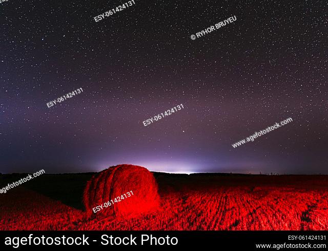 Glowing Stars And Sunset Lights Hay Bales After Harvest. Agricultural Colorful Background Copy Space. Night Starry Sky Above Rural Landscape Field Meadow With...