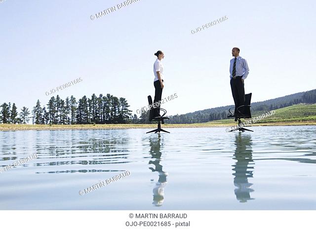 Businesswoman and man standing on office chairs on water