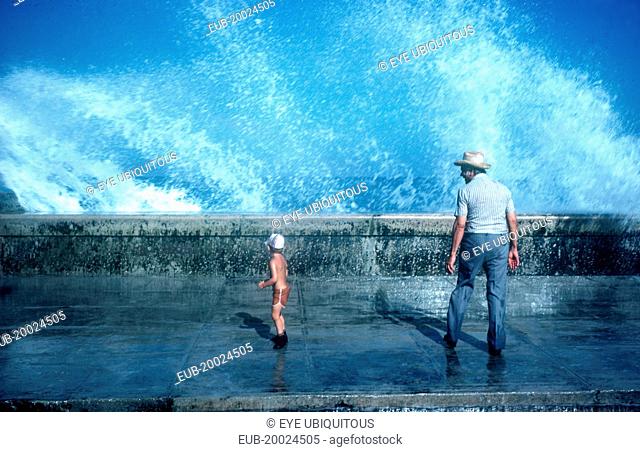 Old man and small boy standing on pavement with waves crashing over the sea wall