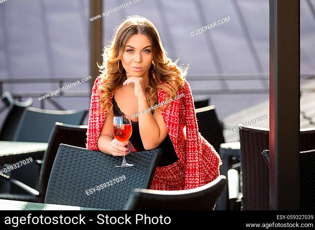 Young fashion woman with glass of wine at sidewalk cafe. Stylish female model in red tweed jacket and shorts suit