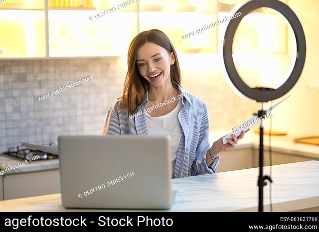 Positive communication. Smiling affable long-haired woman sitting at table talking looking at laptop screen holding papers