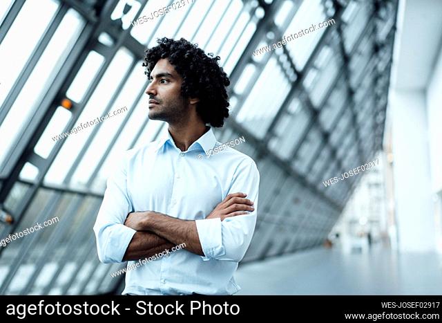 Thoughtful young male professional standing with arms crossed while looking away at workplace