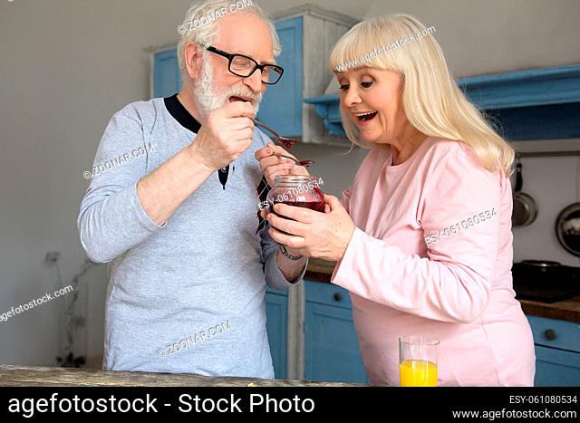 Sweet grandparents trying jam in kitchen. Cute old man and woman dressed in pajamas eating jam straight out of jar in morning