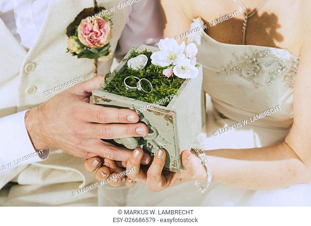 detail shot of a beautifully decorated wooden box with moss and two wedding rings held by hands. In the background one sees bride and groom