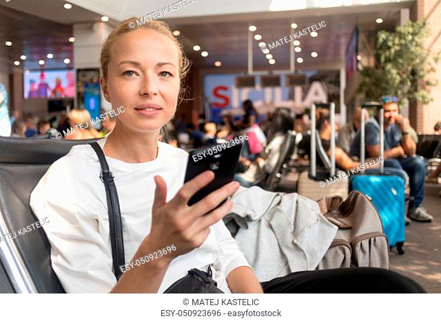 Casual blond young woman reading on her mobile phone while waiting to board a plane at the departure gates at the airport terminal. Travel concept