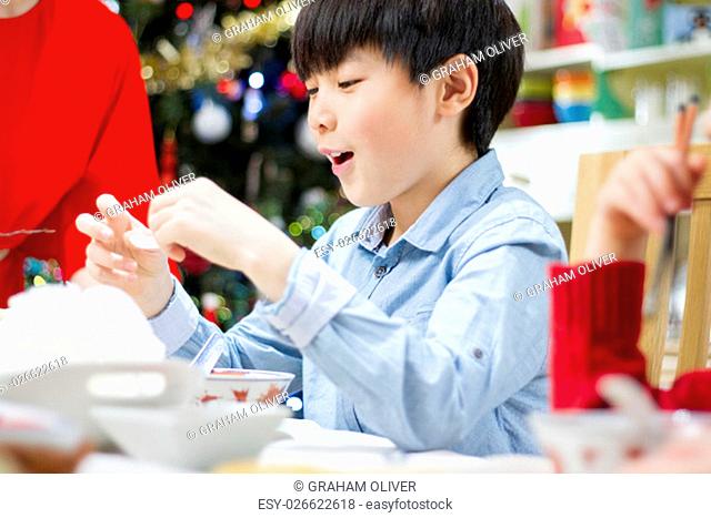 Close up shot of a Chinese boy eating prawn crackers at Christmas dinner with his family. He has a happy expression on his face