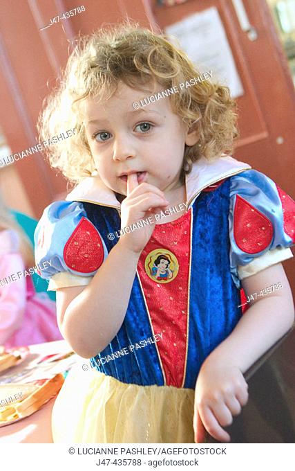 3 year old girl at a party wearing a party dress looking straight into camera, with hand to mouth